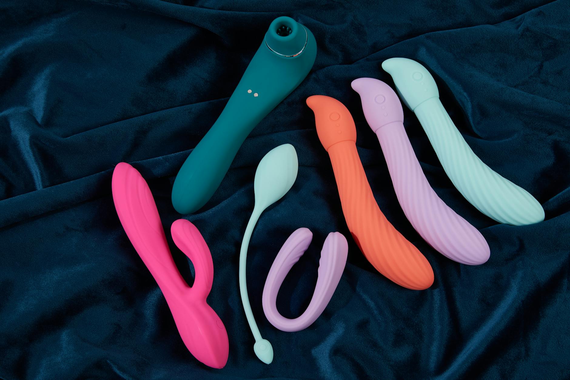 Display di vibratori colorati in silicone su tessuto blu, mostrando forme e colori vivaci per la stimolazione clitoridea.