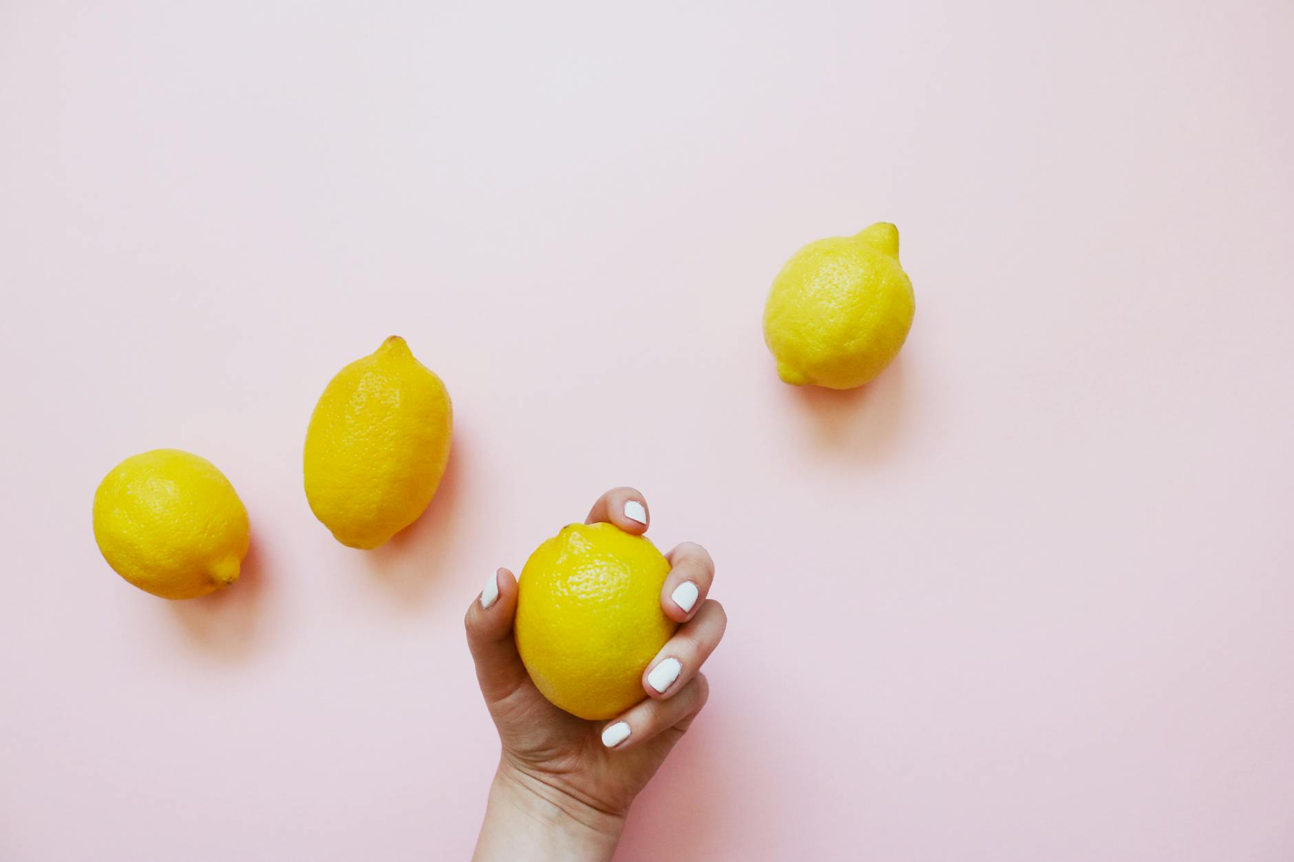Hand holding a yellow lemon on a soft pink background with additional lemons