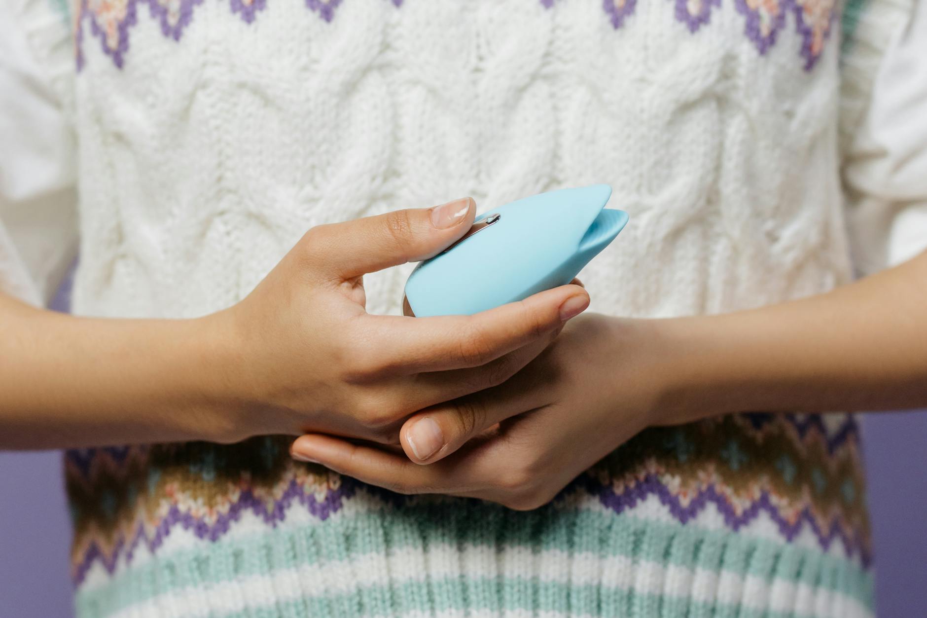 Close-up of hands holding a blue personal massager against a knitted sweater, representing self-care and pleasure exploration